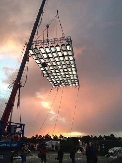 A large flown lighting matrix lifted by crane against a sunset sky at a production site